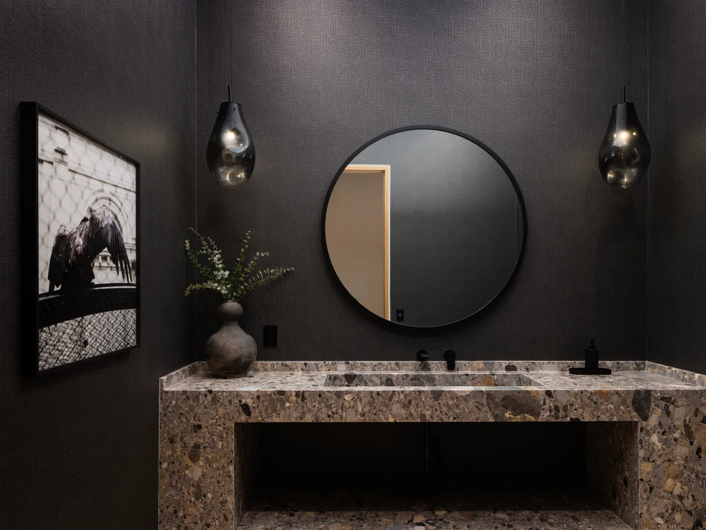 Dark powder room with a broad stone vanity, round mirror and two wall lights.