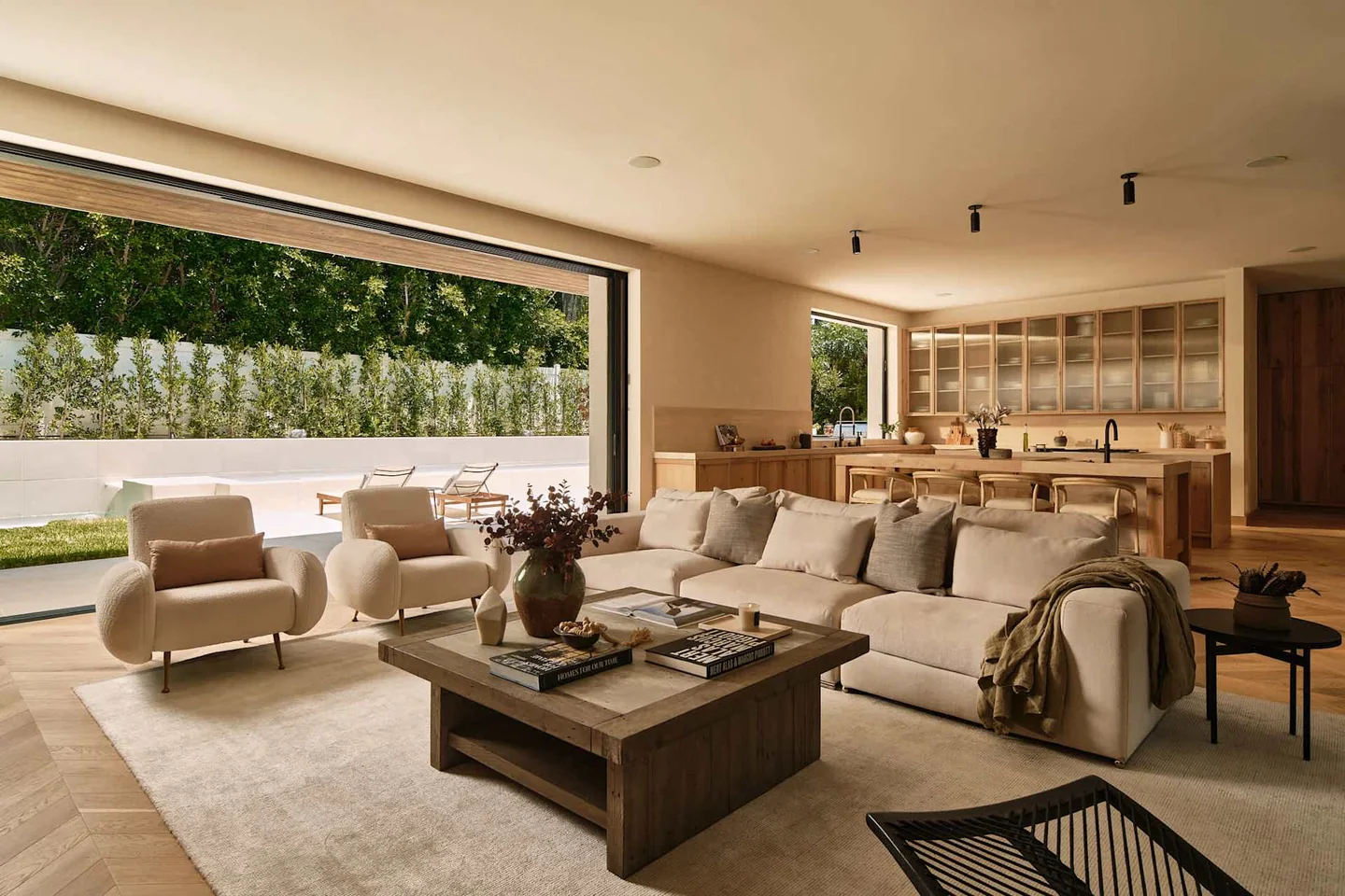 Dobbins living room opening toward a pool garden through broad dark-framed glazing, with timber kitchen cabinetry beyond.