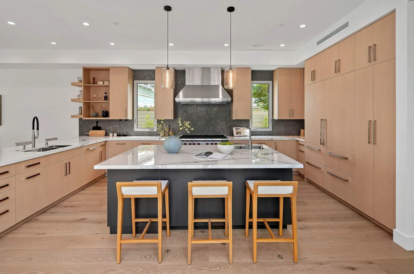 Fairburn kitchen with pale timber cabinetry, a dark island base, pale countertop and three stools beneath slender pendants.